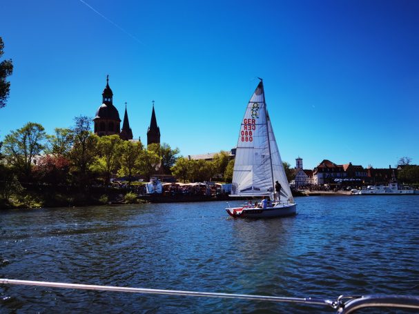 Segelboot auf einem ruhigen Fluss mit historischem Stadtbild im Hintergrund.
