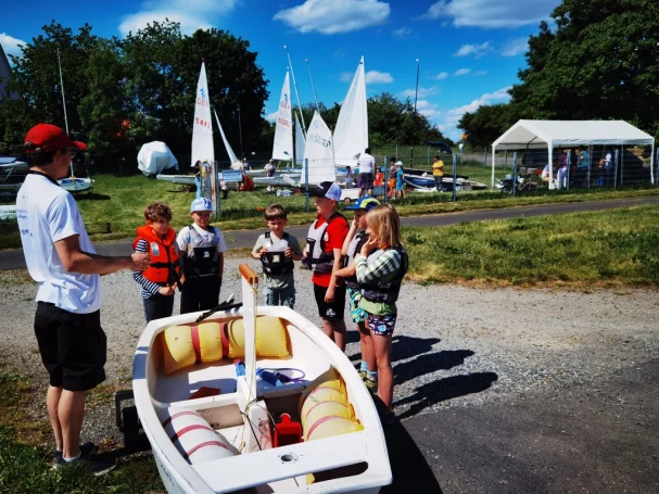 Gruppe von Kindern mit Segelbooten und Trainer am Wasser, an einem sonnigen Tag.